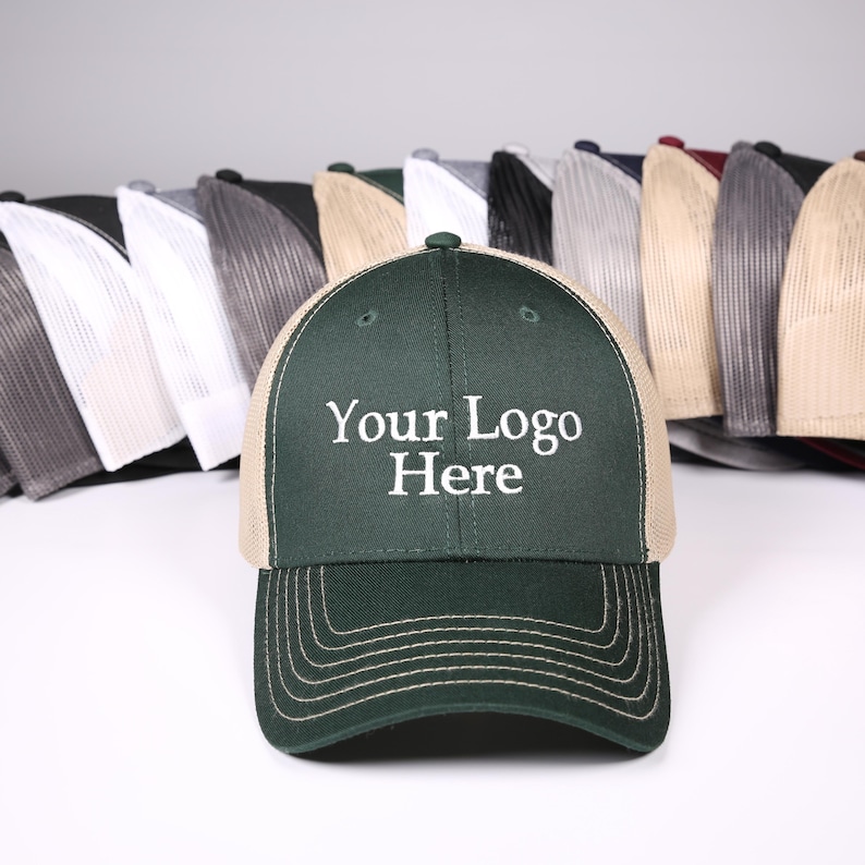 Puede incluir: Gorra de camionero verde oscuro con la parte trasera de malla beige. La parte delantera de la gorra muestra el texto "Your Logo Here" en blanco. Varias gorras m&aacute;s de varios colores son visibles en el fondo.