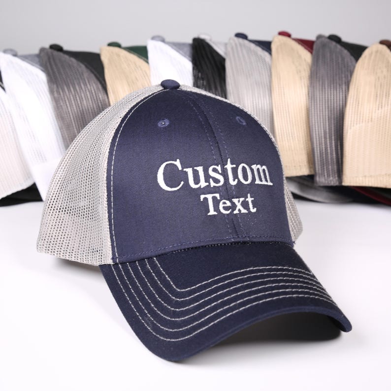 Puede incluir: Gorra de camionero azul marino con la parte trasera de malla blanca y visera curva. El panel frontal muestra las palabras "Custom Text" en blanco. Se ven varias gorras de otros colores en el fondo.