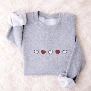 Può includere: Una felpa grigio mélange con girocollo e maniche lunghe. Il design presenta cinque cuori in fila, alternando rosa e rosso, con un cuore bianco al centro. Le maniche sono arrotolate.