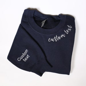 May include: A navy blue sweatshirt with white embroidered text that says "Custom text" on the front and sleeve.
