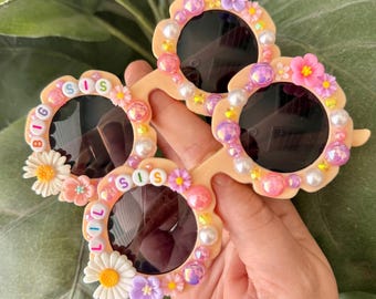 May include: Four pairs of decorative sunglasses with dark lenses. The frames are light peach and adorned with pearls, flowers, and iridescent gems. Two pairs have the word "SIS" spelled out, and one pair says "BIG SIS" and "LIL SIS".
