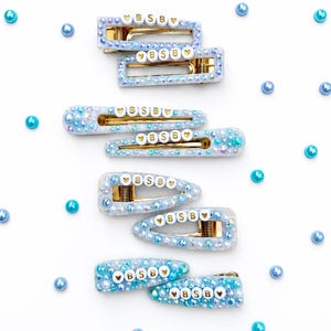 May include: Eight hair clips in various rectangular and triangular shapes, featuring light blue resin embedded with iridescent rhinestones, pearls, and white letter beads spelling BSB flanked by small gold heart icons on each clip.