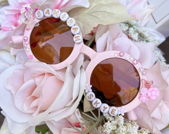 Può includere: Occhiali da sole rotondi rosa chiaro con lenti marroni, decorati con perline bianche con lettere che compongono SAMIRAH sulla montatura sinistra e SKYE su quella destra, arricchiti da piccoli fiori, farfalle e decorazioni simili a perle.