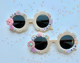 May include: Two pairs of white plastic sunglasses with black lenses. The sunglasses are decorated with flowers and rhinestones. One pair says "BIG SIS" and the other says "LIL SIS".
