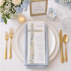 Peut inclure: Une table joliment dressée pour un baptême. Le centre de table est un menu avec un motif de croix florale. Des couverts dorés, une bougie et une petite boîte cadeau avec une croix sont également présents. Le menu indique "God Bless Our Little One BAPTISM".