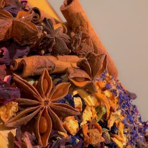 May include: Close-up of a wooden surface with a variety of dried herbs and spices. Star anise, cinnamon sticks, and dried flowers in shades of brown, orange, blue, and purple are visible. The image suggests a tea blend or spice mix.