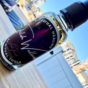 May include: A clear glass spray bottle with a black cap, filled with a golden liquid. The label reads "Shiloh Wellness Spiritual Healing Tools" in white script and text. The bottle is on a wooden surface, with a blue sky and buildings in the background.