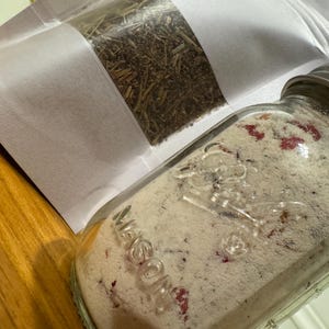 May include: A clear glass Mason jar filled with a light-colored powder containing small red flecks, paired with a white stand-up pouch containing dried brown botanical herbs. The jar features a silver metal lid and embossed glass lettering.