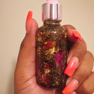 May include: A clear glass bottle with a pink dropper top filled with a golden liquid and dried flowers and gold flakes.