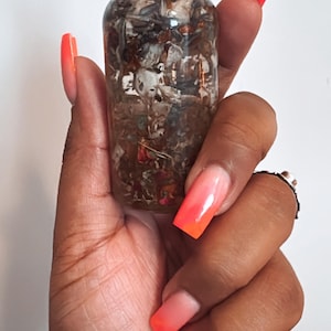 May include: A clear glass bottle with a black dropper top filled with a mixture of dried herbs and flowers. The bottle is being held by a hand with orange nail polish.
