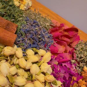 May include: A wooden surface displays an assortment of dried botanicals. The arrangement includes yellow jasmine flowers, blue lavender buds, pink rose petals, cinnamon sticks, and green herbs. The composition is a colorful display of natural elements.