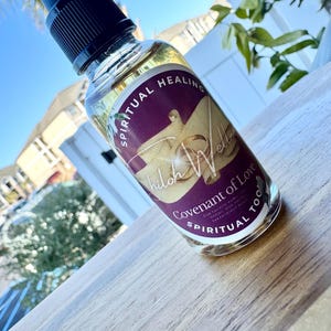 May include: A clear glass spray bottle with a black cap, filled with a yellow liquid. The bottle has a label with the words "Spiritual Healing", "Shiloh Wellness", and "Covenant of Love". The background is a wooden surface and a blurred outdoor scene.