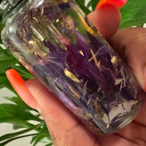 May include: A clear glass bottle filled with a purple and yellow liquid and dried purple flowers. The bottle is held in a hand with bright orange nail polish.