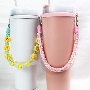 May include: Two reusable tumblers with colourful crocheted handles. The tumbler on the left is white with a yellow and blue flower design. The tumbler on the right is pink with a white and pink flower design.