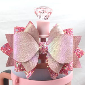 Peut inclure: Un nœud rose et pailleté iridescent avec un dessus en strass transparent, attaché à un gobelet rose.