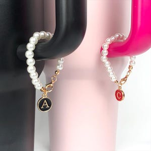 Pode incluir: Duas pulseiras de pérolas com pingentes de ouro. Um pingente é preto com a letra A e o outro é vermelho com a letra C.
