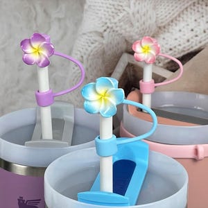 Könnte beinhalten: Drei Edelstahlbecher mit unterschiedlichfarbigen Deckeln und floralen Strohhalm-Toppern. Die Becher sind mit Wasser gefüllt und die Strohhalme werden mit einem Silikonband befestigt.