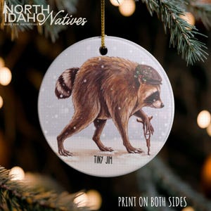 May include: A round white ceramic ornament featuring a brown raccoon wearing a small cap with holly, walking with a crutch in the snow. The text "TINY JIM" is printed below the raccoon. A gold string is attached to the top for hanging.