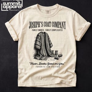 May include: Cream-colored t-shirt with rolled sleeves featuring a black and white illustration of a coat and spools of thread. The text "JOSEPH'S COAT COMPANY" and "Favor Looks Good on You" are printed on the shirt.