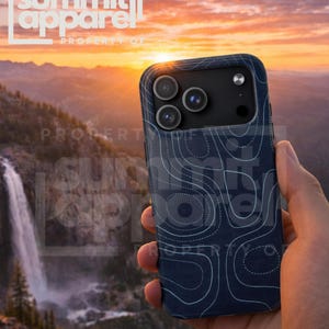 May include: A navy blue phone case with a white topographic map design is held in a hand. The case has a raised edge around the camera lenses. The background shows a scenic mountain landscape with a waterfall and sunset.
