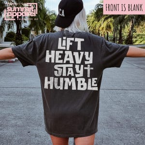 May include: Dark grey t-shirt with the text "LIFT HEAVY STAY HUMBLE" in white block letters. The shirt has a vintage, worn appearance. The person in the image is wearing a black baseball cap with "CA" on it.