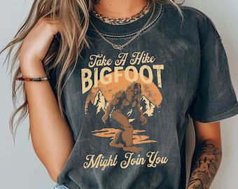 May include: A black t-shirt with a vintage-style graphic of a Bigfoot walking through a forest. The text on the shirt reads "Take A Hike Bigfoot Might Join You".