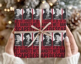 May include: A wrapped gift with a festive design. The wrapping paper features mugshots of two men with the text "Wet Bandits" and "Escaped". The gift is tied with a cream-coloured ribbon and bow.