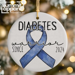 Op de afbeelding: Ronde witte keramische ornament met een blauw lint en de tekst "DIABETES warrior SINCE 2024". Het heeft een gouden ophangtouwtje. De achtergrond is wazig met bokeh-lichten.