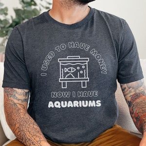 May include: A dark grey t-shirt with a white graphic that reads "I used to have money now I have aquariums". The graphic includes a simple line drawing of a fish tank with a fish inside.