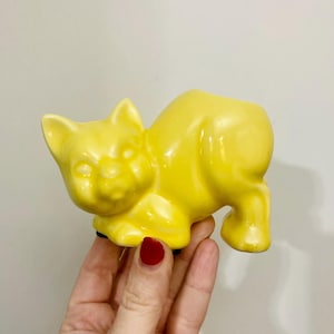 May include: A bright yellow ceramic cat figurine. The cat is in a playful pose, with its body arched and legs extended. The figurine has a glossy finish and is a solid, cheerful yellow color. The cat's features are detailed, including its ears, eyes, and nose.