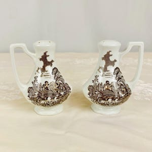 May include: A pair of white ceramic salt and pepper shakers with a brown transferware design. Each shaker has a handle and a detailed landscape scene. The design includes trees, buildings, and a bridge. The shakers have a classic, vintage aesthetic.