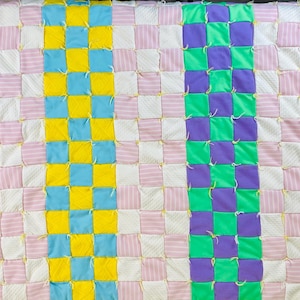 May include: A colorful woven quilt with a patchwork design. The quilt features squares of various colors, including purple, green, yellow, blue, pink, and white. The squares are arranged in a grid pattern, creating a visually appealing and textured surface.