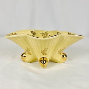 May include: A pale yellow, scalloped-edge ceramic dish with gold floral accents and three gold ball feet. The dish has a fluted design and a gold trim around the rim. The gold floral pattern is repeated on the dish's exterior.