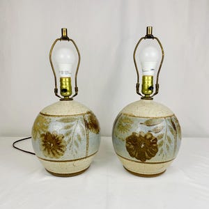 May include: Pair of ceramic table lamps. Each lamp has a round base with a floral design in brown and gold on a light blue and beige background. The lamps have brass-colored hardware and a white light bulb.