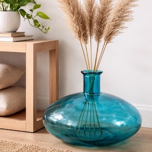 May include: A large, turquoise glass vase with a wide, rounded base and a narrow neck. The vase holds several tall, dried, beige-colored ornamental grasses. The vase sits on a wooden floor next to a woven rug.