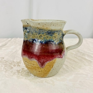May include: A handmade ceramic pitcher with a cream-colored handle and spout. The pitcher features a textured surface with bands of color, including red, yellow, blue, and brown. The pitcher is a unique piece of pottery.
