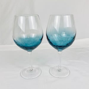May include: Two clear glass wine glasses featuring a distinct crackle texture at the base of the bowl. The lower portion of each bowl displays a vibrant teal blue gradient that fades into clear glass toward the top rim, supported by a stem and base.