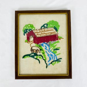 May include: Embroidered artwork in a brown wooden frame. The image depicts a red barn with a brown roof, a blue waterfall, green trees, and a sheep. The artwork is on a beige fabric background.