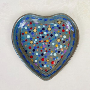 May include: A heart-shaped ceramic dish with a speckled design. The dish features a blue rim with a dotted pattern and a gray interior filled with colorful polka dots in red, blue, yellow, and white. The dish is on a white surface.