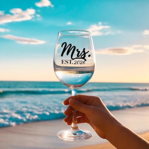 Puede incluir: Una copa de vino transparente llena de líquido, sostenida contra un fondo de playa. La copa tiene la palabra "Mrs." y "EST. 2026" en escritura negra. El fondo presenta un cielo azul, el océano y arena.