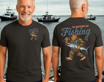 May include: Dark gray t-shirt with a Bigfoot graphic carrying a marlin and fishing rod. The text "I'd Rather Be Fishing" is printed on the front and back of the shirt. The shirt is worn by a person in front of a body of water with boats.