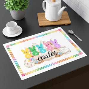 Puede incluir: Mantel individual blanco con un borde de arcoíris, con gráficos temáticos de Pascua. El diseño incluye formas de conejito en amarillo, verde, azul, rosa y morado, con la palabra "Easter" en cursiva. También hay huevos decorativos.