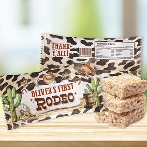 May include: A brown and white cow print treat bag with the text "Oliver's First Rodeo" and a cactus illustration. The bag is filled with a stack of three white rice crispy treats.