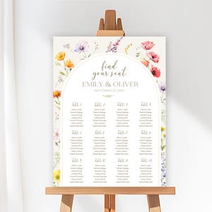 Könnte beinhalten: Ein Hochzeits-Sitzplan auf einer Staffelei. Das cremefarbene Schild zeigt florale Aquarellakzente und den Text "find your seat" über den Namen des Paares und dem Datum. Die Namen der Gäste sind unter den Tischnummern aufgeführt.