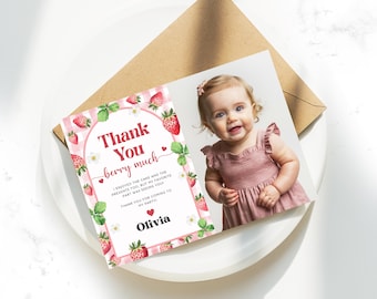 May include: A thank you card with a photo of a baby girl in a pink dress. The card has a strawberry theme with the text "Thank You berry much" and the name "Olivia". The card is on a white plate with a brown envelope.