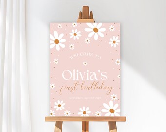 Op de afbeelding: Een roze bord met witte madeliefjes en de tekst "Welcome to Olivia's first birthday" in witte en gouden letters. Het bord wordt gepresenteerd op een houten ezel, perfect voor een feest.
