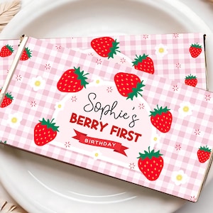 Peut inclure: Deux boîtes rectangulaires avec un motif vichy rose, ornées de fraises rouges et de fleurs blanches. Les boîtes présentent le texte "Sophie's Berry First Birthday", parfaites pour une première fête d'anniversaire.