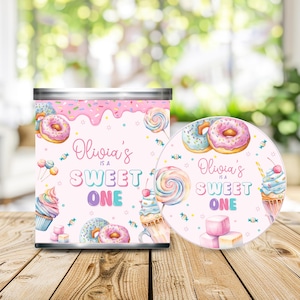 May include: A cylindrical metal container and a circular lid featuring a pink background with illustrations of donuts, cupcakes, lollipops, and candy. The text reads Olivia's IS A SWEET ONE in pink and blue lettering with a pink frosting drip design.