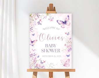 May include: A baby shower welcome sign on an easel. The sign features a watercolor design with butterflies and floral elements in shades of purple and pink. The text reads "Welcome to Olivia's Baby Shower" and the date "September 25, 2025".