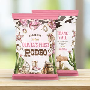 May include: Two pink-topped party favor bags with a rodeo theme. The bags feature illustrations of a cowboy hat, boots, cactus, and the text "Olivia's First Rodeo". The bags also say "Thank Y'all".